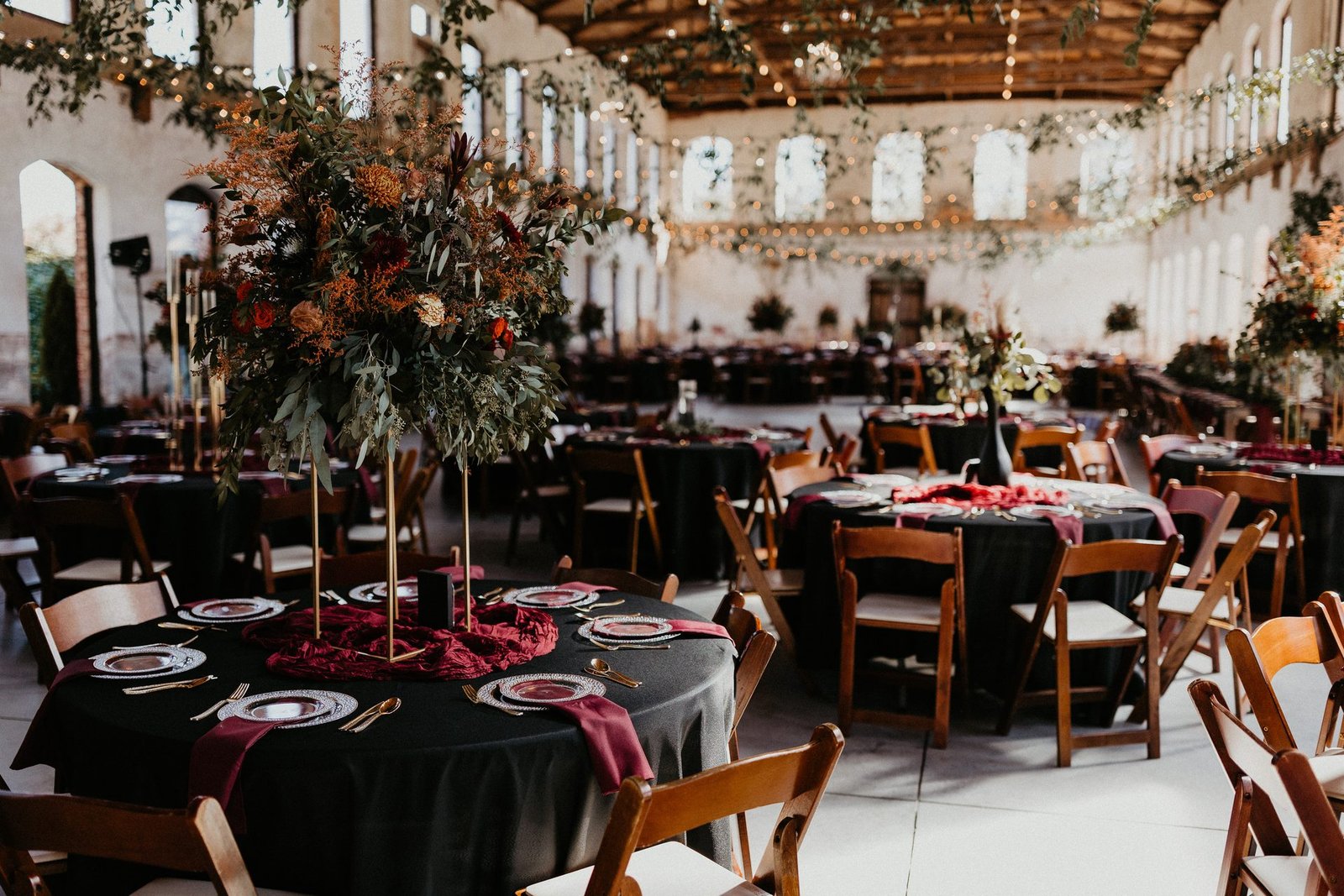 Reception hall with black-linen round tables, tall autumn floral centerpieces and warm string lights