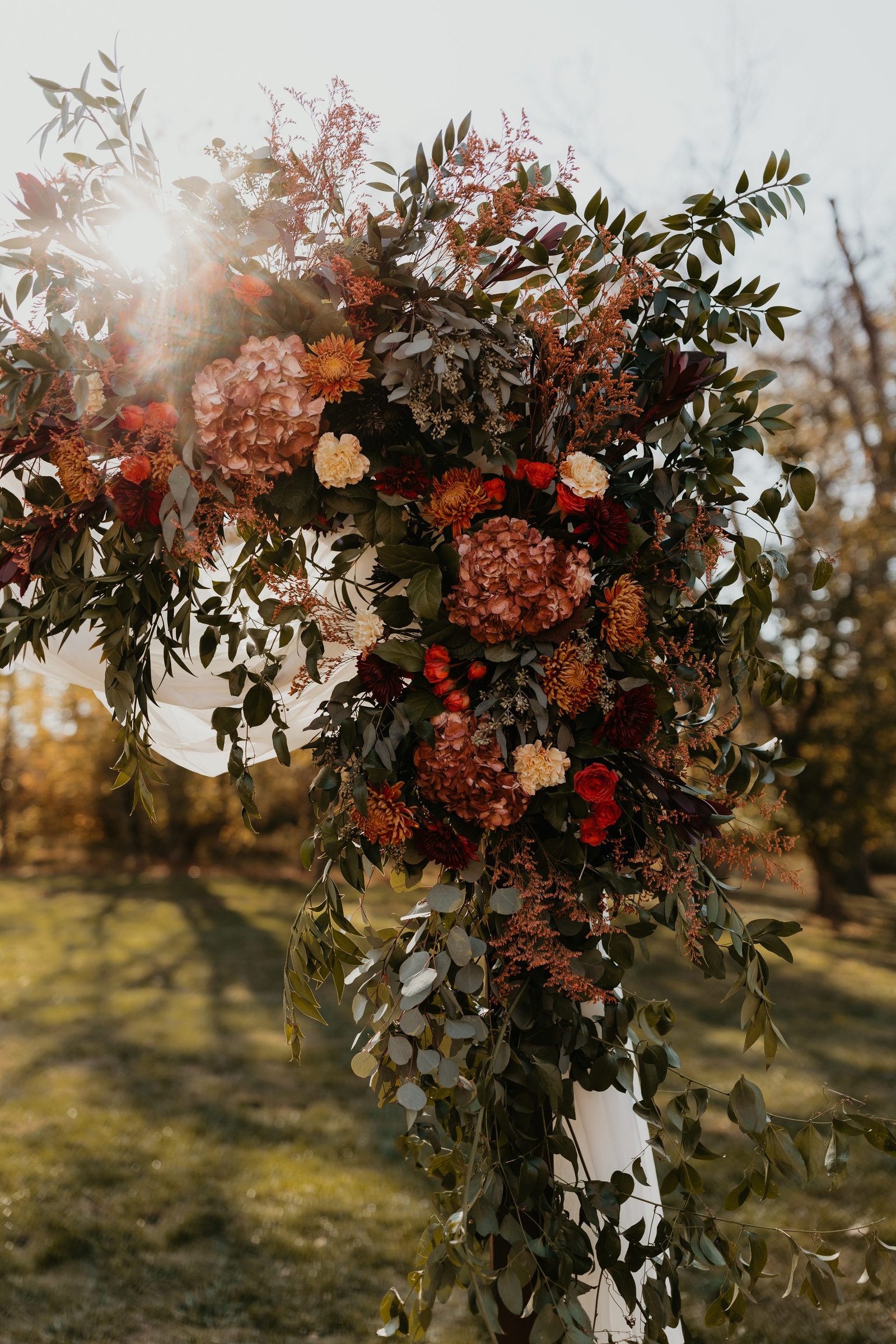 Autumn ceremony arch of rust, terracotta and burgundy blooms with eucalyptus, backlit at golden hour